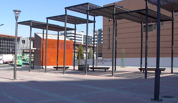 espacio urbano contemporáneo caracterizado por una serie de pérgolas metálicas de color oscuro que crean zonas de sombra sobre bancos lineales